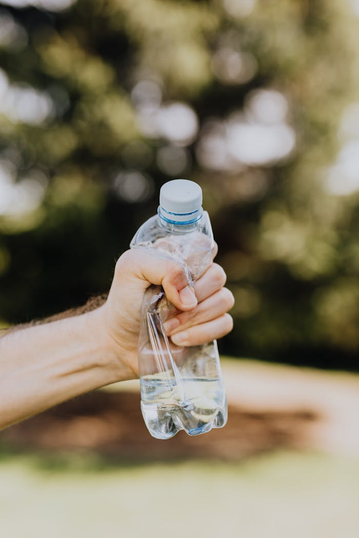 our-story Crop unrecognizable man crumpling plastic bottle of water in hand against blurred background of green park trees on sunny summer day